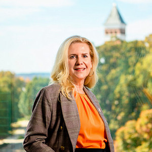 A portrait of a woman in a stylish outfit standing by a large window with a scenic view of trees and a tower in the background.