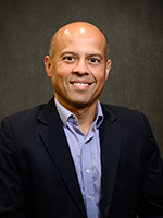 Professional headshot of a man with a shaved head, wearing a navy blazer over a light purple shirt, smiling at the camera.