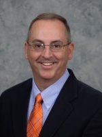 Professional headshot of a man in a blazer and orange tie, smiling against a textured gray background.