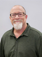 Headshot of a man with glasses and a beard, wearing a green collared shirt, smiling against a neutral background.