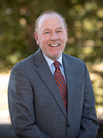A smiling man in a suit and tie, standing outdoors with a blurred green background.