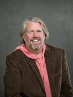 A smiling man with long hair wearing a brown blazer over a pink hoodie.