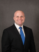 Professional headshot of a smiling man in a suit with a blue tie against a dark background.