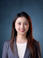 Professional headshot of a woman with long dark hair, wearing a blazer and smiling against a textured blue background.