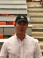 A man wearing a black cap with "LA" embroidered on it, standing in a lecture hall with orange seating.