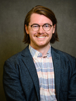 Headshot of a smiling man with glasses and long hair, wearing a plaid shirt and a blazer.