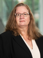 Professional headshot of a woman with long, brown hair wearing glasses and a black blazer.