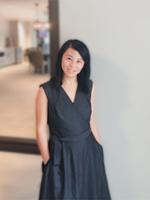 A smiling woman in a dark dress stands confidently with her hands in her pockets against a softly blurred interior background.