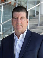 Professional headshot of a man wearing a suit and smiling, set against a modern architectural background.