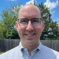 A smiling man wearing glasses and a button-up shirt in a sunny outdoor setting.