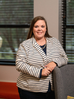 A professional portrait of a woman in a striped blazer, standing beside a gray partition in a well-lit office setting.