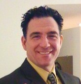 A professional man with dark hair wearing a suit and tie, smiling confidently.