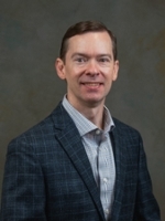 Professional headshot of a man in a blazer, smiling against a textured background.