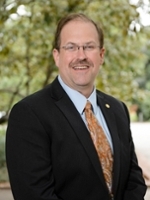 Professional portrait of a smiling man in a suit and tie, set against a blurred outdoor background.