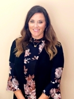 A woman with long brown hair wearing a floral blouse and black vest smiles in front of a light-colored background.