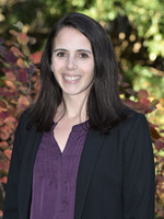 A professional headshot of a woman with long dark hair, wearing a purple blouse and black blazer, set against a backdrop of colorful autumn foliage.