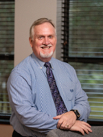 A smiling man in a blue dress shirt and a patterned tie, posing confidently in a well-lit office environment.