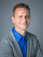Professional headshot of a man wearing a blue shirt and gray blazer, smiling confidently.
