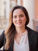 Professional headshot of a woman with long hair wearing a black blazer and white blouse, smiling confidently.