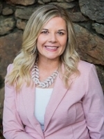 A professional headshot of a woman with blonde hair, wearing a pink blazer and a statement necklace, set against a natural stone background.