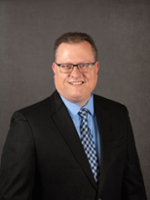 Professional headshot of a man wearing glasses, a black suit, and a blue shirt with a patterned tie.