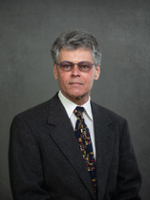 Professional portrait of a middle-aged man with gray hair wearing glasses and a suit, set against a neutral background.