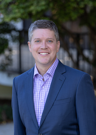 Professional portrait of a smiling man in a suit, set against a blurred outdoor background.