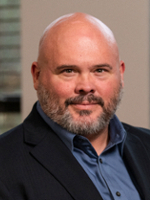 Professional headshot of a man with a beard, wearing a blue shirt and dark blazer.