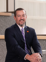 Professional portrait of a man in a suit sitting on stairs, smiling, with a purple tie and an orange logo on his lapel.
