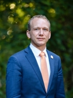 A professional portrait of a man in a blue suit and orange tie, standing outdoors against a blurred green background.
