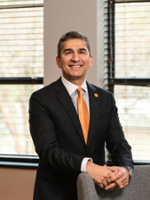 A professional portrait of a smiling man in a black suit and orange tie, standing beside a chair in an office setting.