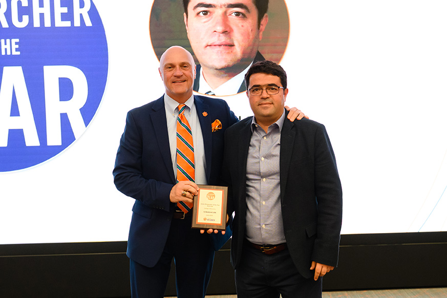 Two men stand together, one holding a plaque and smiling, during an awards ceremony. The background features a large display with the text "Researcher of the Year."