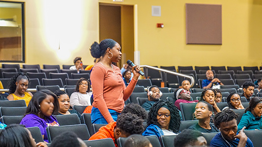 Young person of color holding a microphone in an auditorium surrounded by a group of her peers looking at her as she speaks.