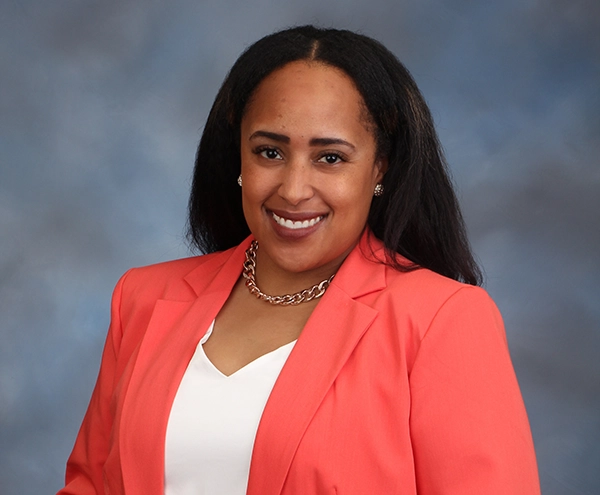 Professional headshot of a woman wearing a coral blazer and smiling against a soft blue background.