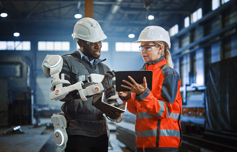 Man in ergonomic suit looking at  tablet with female coworker.