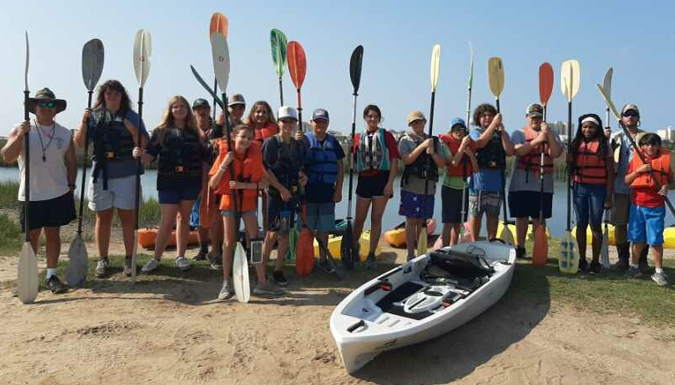 group of students on the shore holding paddles and  standing in front of a kayak