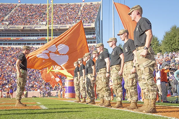 A row of Clemson cadets stand at attention in Memorial Stadium beside waving orange Tiger Paw flags and fan-filled seats. 