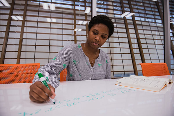 Female student writes on dry erase table next to an open book.