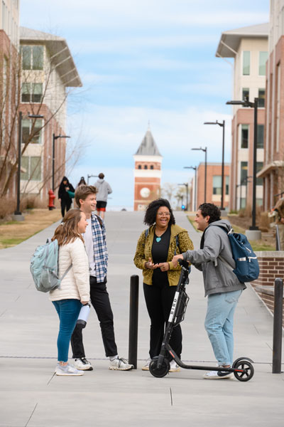 A male student with a mobile scooter talks with another male student and two female students on a concrete walkway on campus. 
