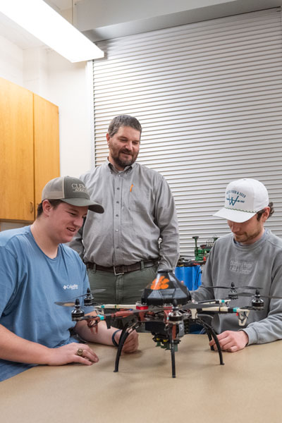 Two male students sit at a table with a drone device as a male professor stands over them and looks on. 
