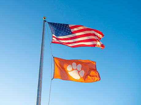 Flag pole with American flag and an orange flag with a white tiger paw representing Clemson University blowing in the wind. 