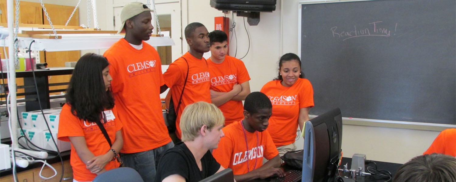 Students in orange shirts looking at a computer screen.