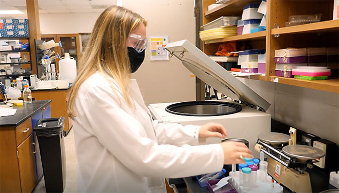 Female student working in lab, wearing masks. Frame from video.