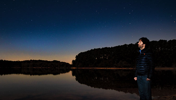 Man (Marco Ajello) on side of lake gazing at star-filled sky.