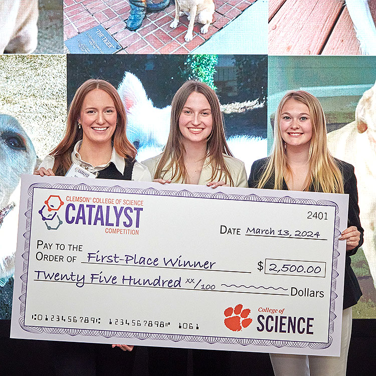 Three females in front of a video wall, holding check.