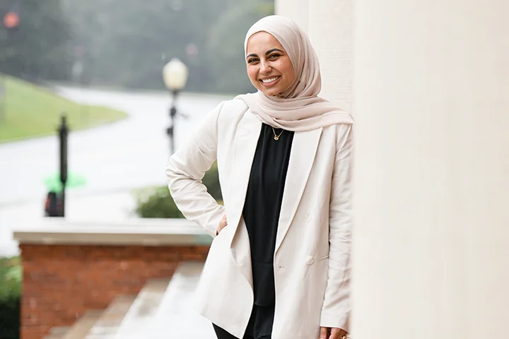 A woman in a white coat and a matching hijab smiles and poses on steps overlooking a nearby road.