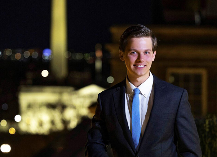 A male student wearing a navy suit, white shirt and royal blue tie stands outside at night. 