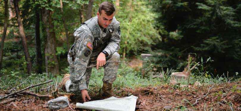 A male Clemson ROTC cadet wearing camouflage kneels in the woods and examines a map. 