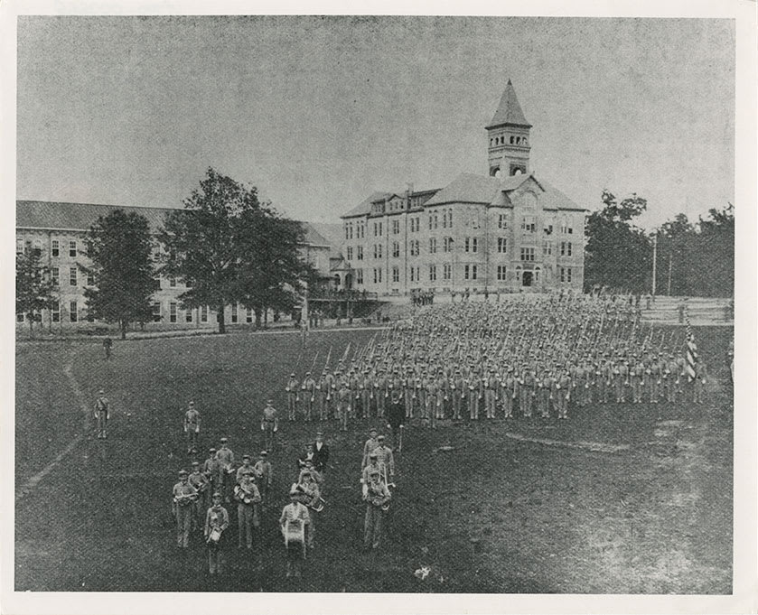 A historic black and white image shows rows of Clemson cadets and a small band performing drills in front of Tillman Hall.