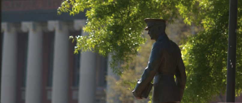 A statue of a Clemson cadet strolling onto Bowman with a book in hand is framed by green trees and the columns of Sikes Hall.